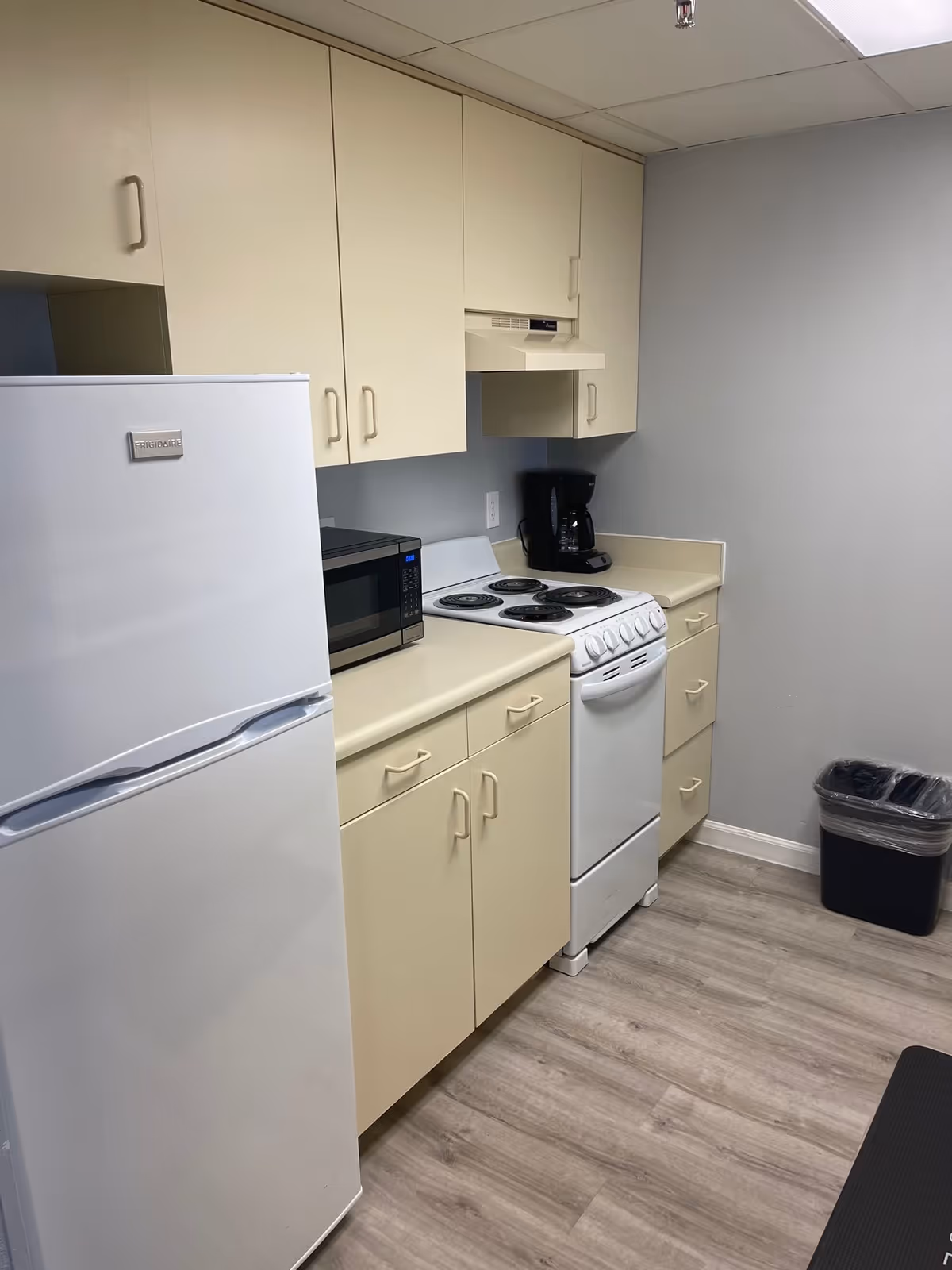 A small kitchen area with beige cabinets, a white refrigerator, a microwave, a white electric stove with four burners, a coffee maker, and a trash bin. The floor is light wood, and the walls are painted light gray.
