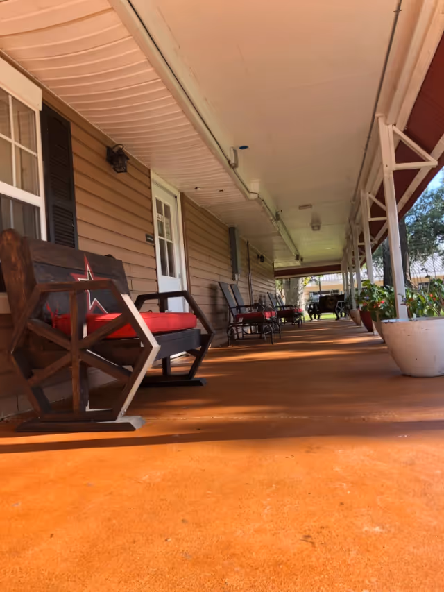 A covered porch area with wooden rocking chairs featuring red cushions and a star design on the backrest. The porch has a brown floor and beige siding on the building wall, with potted plants along the edge and a white ceiling overhead.