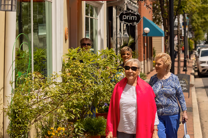 Four elderly women walking along a sunny sidewalk lined with plants and storefronts, with one woman wearing a bright pink shawl and sunglasses.