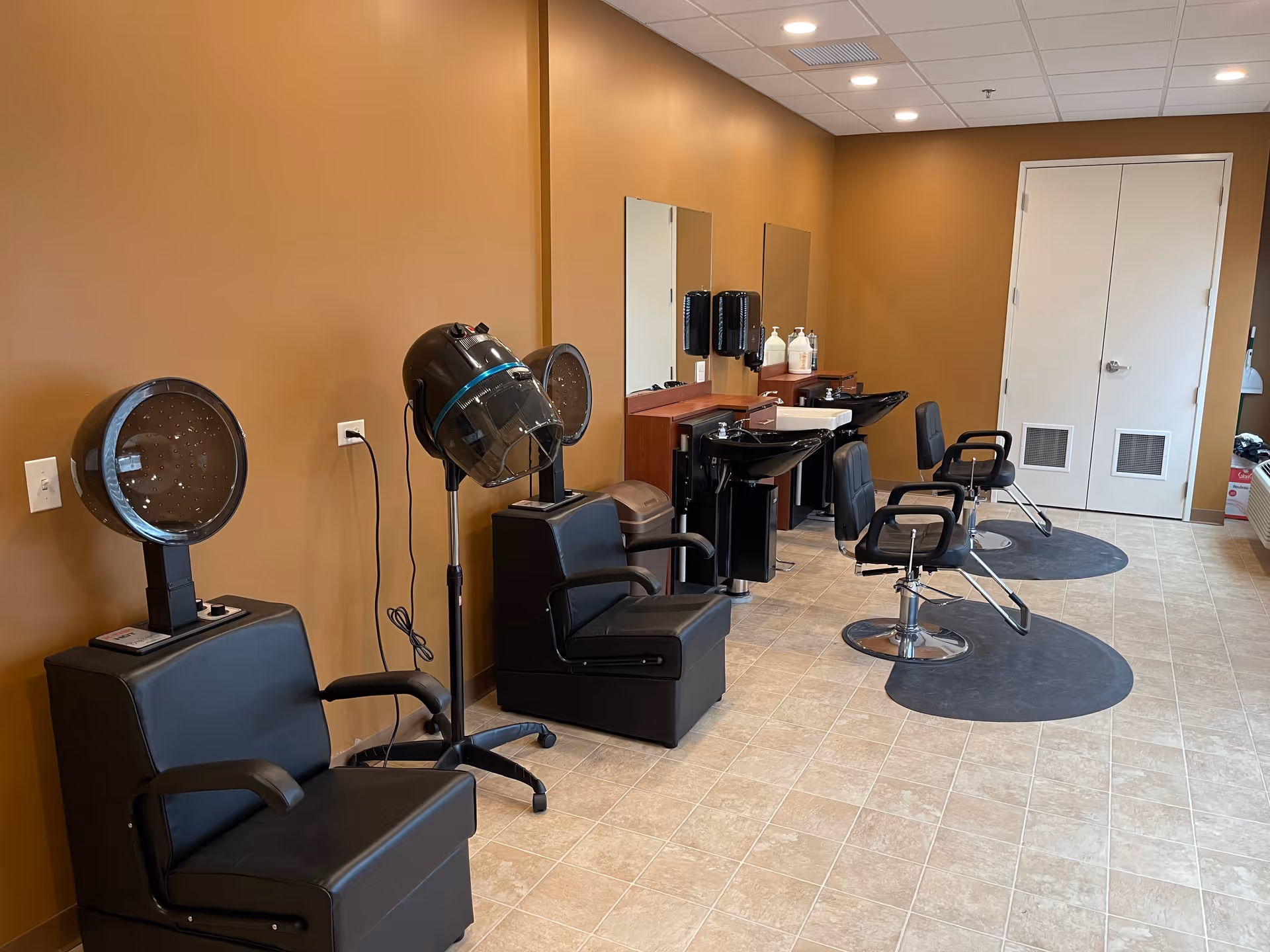 Interior view of a hair salon area with brown walls and tiled floor. The room features two black hair drying chairs with hooded dryers, two black salon chairs in front of sinks for washing hair, mirrors mounted on the wall, and various hair care products on the counter. A double white door is visible at the far end of the room.