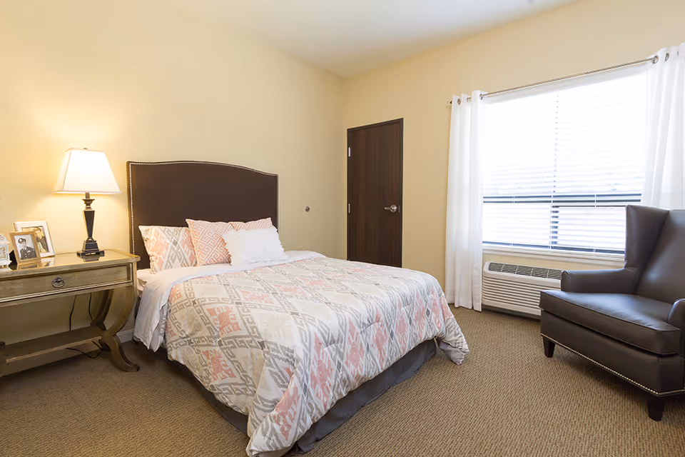 A cozy bedroom in a senior living facility featuring a bed with patterned bedding and multiple pillows, a bedside table with a lamp and framed photos, a dark brown armchair, a window with white curtains and blinds, and a closed wooden door.