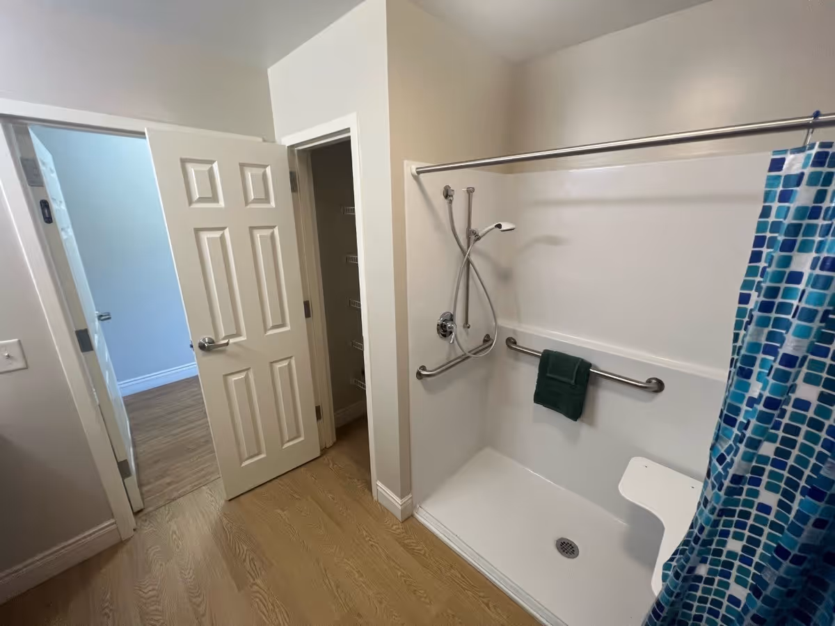 A bathroom with a walk-in shower featuring a handheld showerhead, grab bars, a green towel hanging on one grab bar, and a white fold-down shower seat. The shower has a blue and white patterned curtain. The bathroom floor is wood-style, and there are two open doors leading to adjacent rooms.