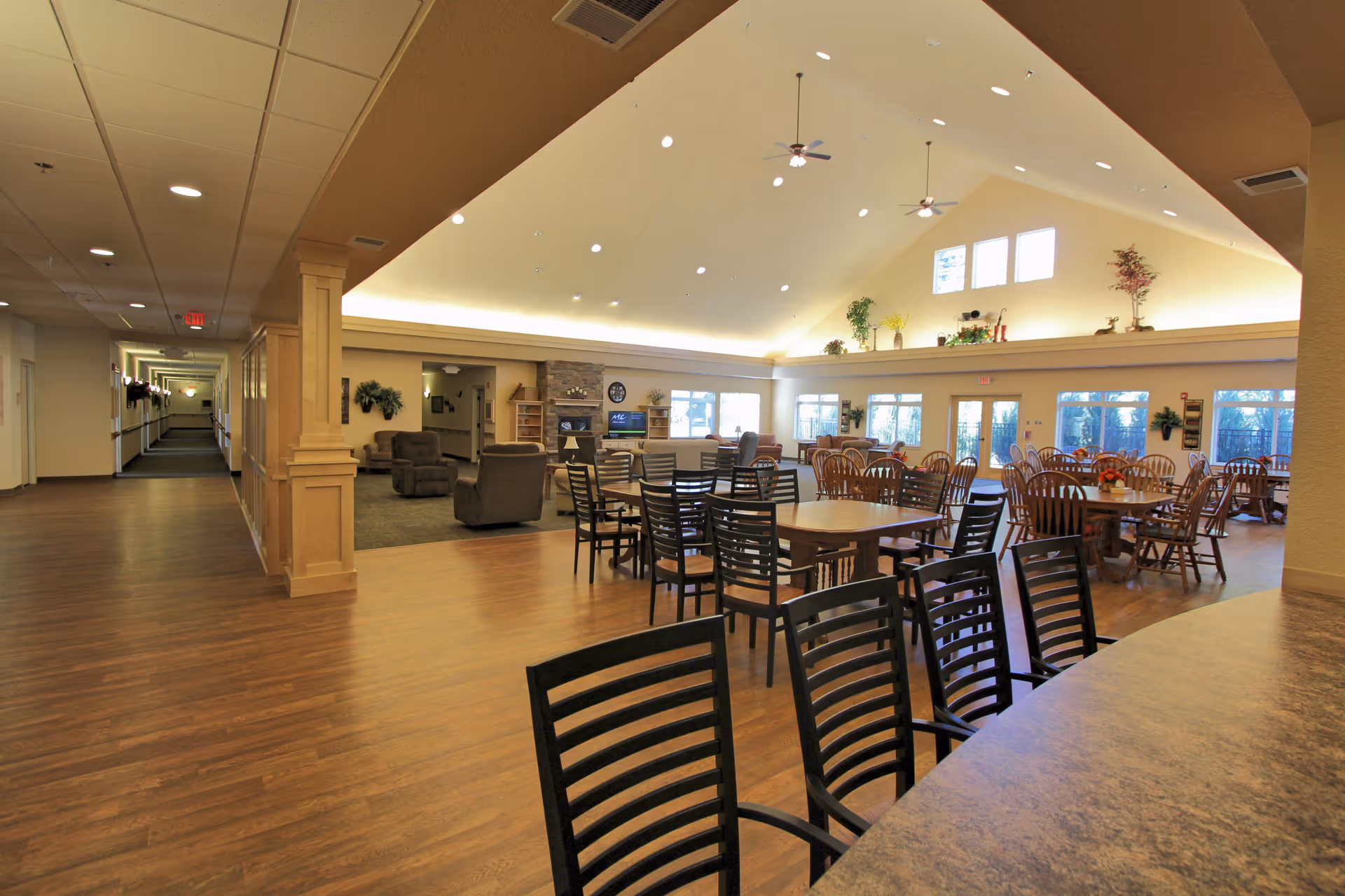 Spacious and well-lit common area in an assisted living facility featuring multiple dining tables with chairs, a seating area with armchairs near a fireplace and television, large windows allowing natural light, and decorative plants on a high ledge under a vaulted ceiling with ceiling fans.