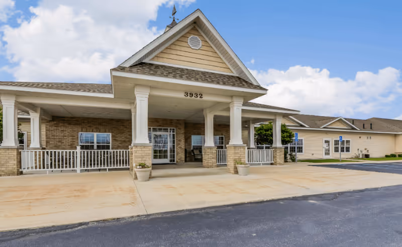 Front exterior view of a single-story assisted living and memory care facility with a covered entrance supported by white columns, beige siding, brick accents, and the number 3932 displayed above the entrance. There is a paved driveway and parking area in front, with clear skies and some clouds in the background.
