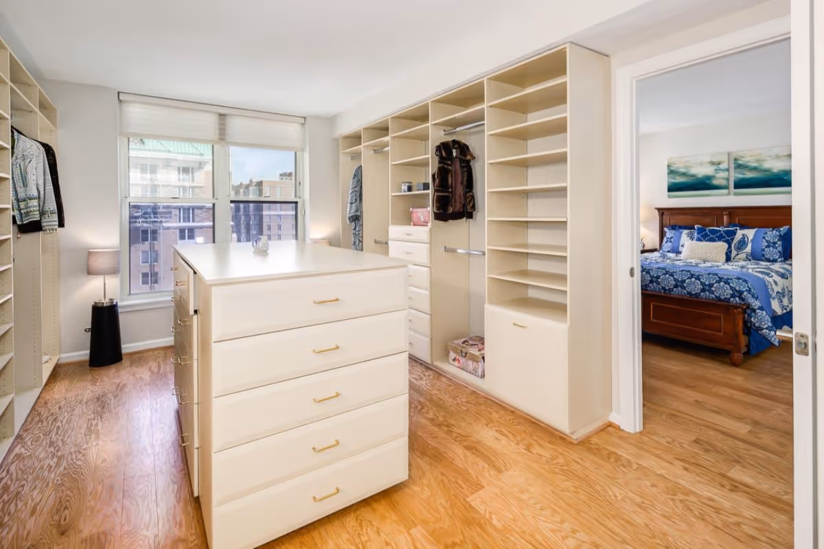 A spacious walk-in closet with built-in shelves, hanging rods, and a central island with drawers. The closet has wooden flooring and a large window letting in natural light. Through an open door, a bedroom with a wooden bed frame and blue bedding is visible.