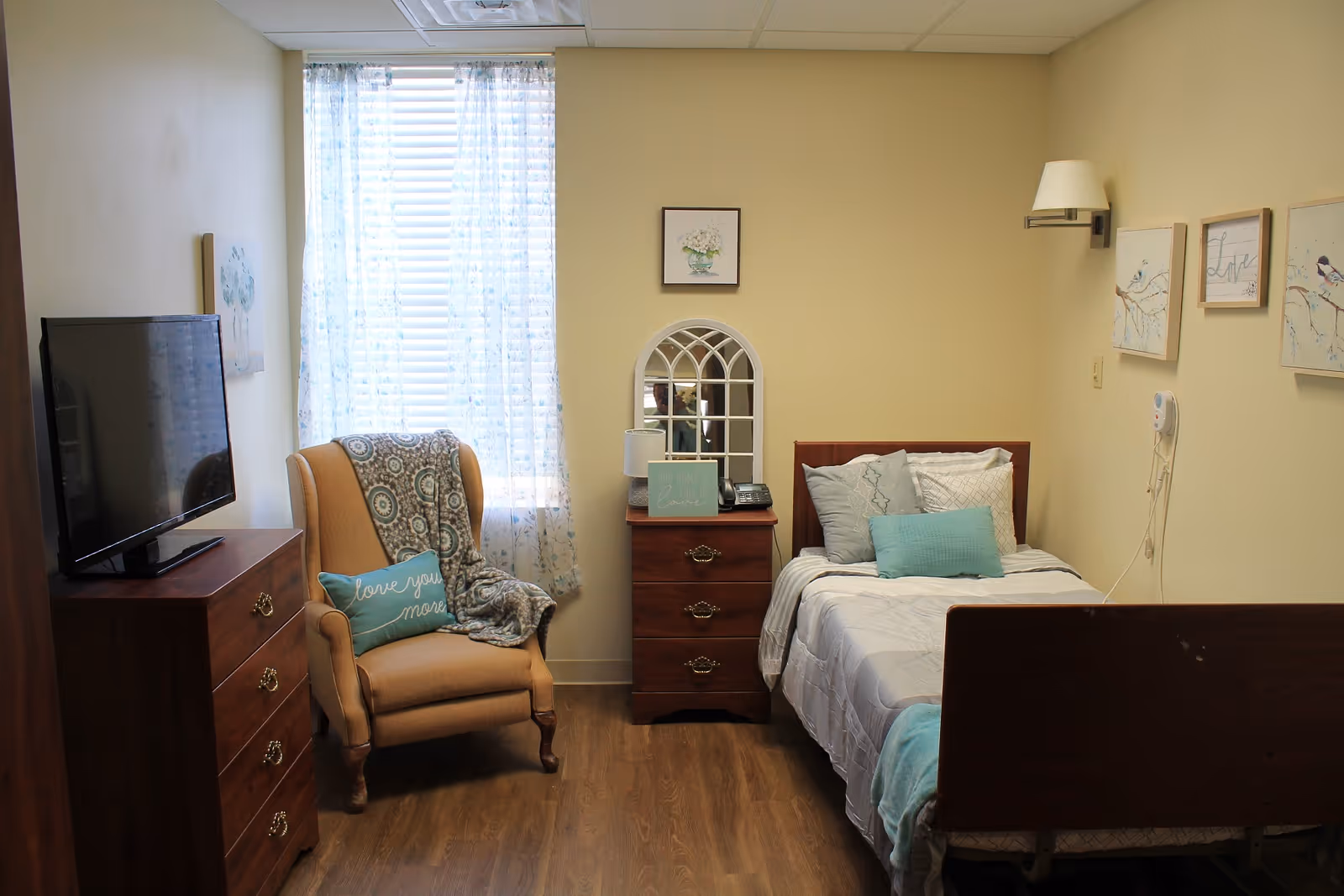 A cozy bedroom in a senior living facility with a single bed made up with white and light blue bedding, a wooden nightstand with a lamp and a decorative mirror, a comfortable armchair with a patterned throw and a pillow that says 'love you more', a wooden dresser with a flat-screen TV on top, and light yellow walls adorned with framed artwork.