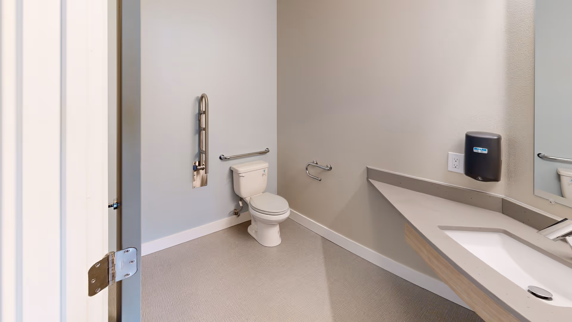 Accessible bathroom with a toilet, wall-mounted grab bars, a long sink countertop and a paper towel dispenser.