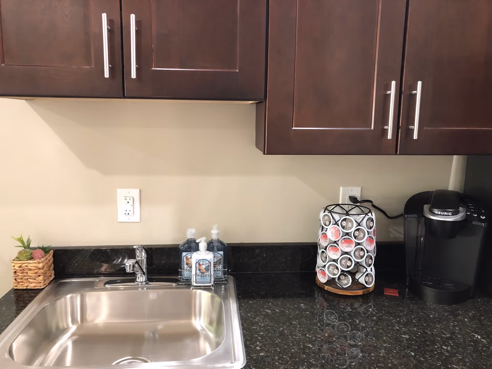 A kitchenette area with a stainless steel sink, dark granite countertop, and dark wooden cabinets. On the countertop, there is a small basket with a plant, two bottles of hand soap, a rotating coffee pod holder filled with coffee pods, and a black Keurig coffee maker.