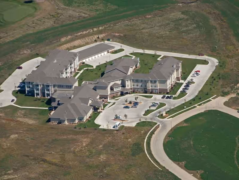 Aerial view of a multi-wing senior living building with driveways, parking areas, and surrounding grassy fields.