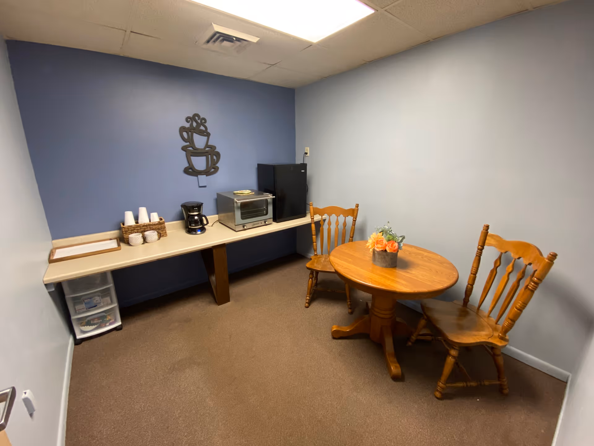 Small room with a wooden round table and two wooden chairs. On the table is a small flower arrangement. Against the wall is a long counter holding a coffee maker, a toaster oven, a small refrigerator, and some cups. The walls are painted light blue and blue, and there is a decorative metal wall art of stacked coffee cups above the counter.