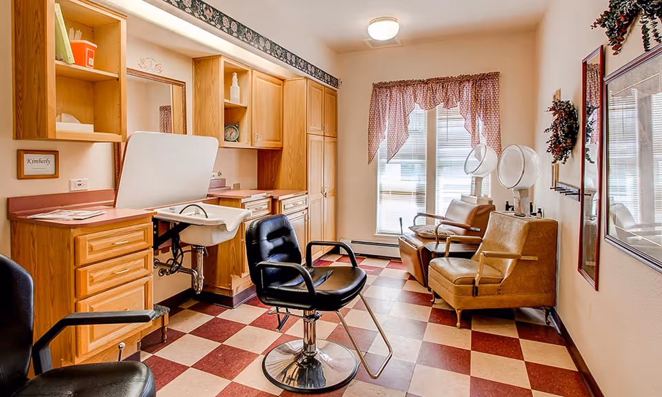 Interior view of a salon area in an assisted living facility with a black salon chair in the foreground, wooden cabinets and a sink with a white basin on the left, two brown leather chairs with hair dryers in front of a window with red and white checkered curtains, and framed pictures and wreaths on the walls.
