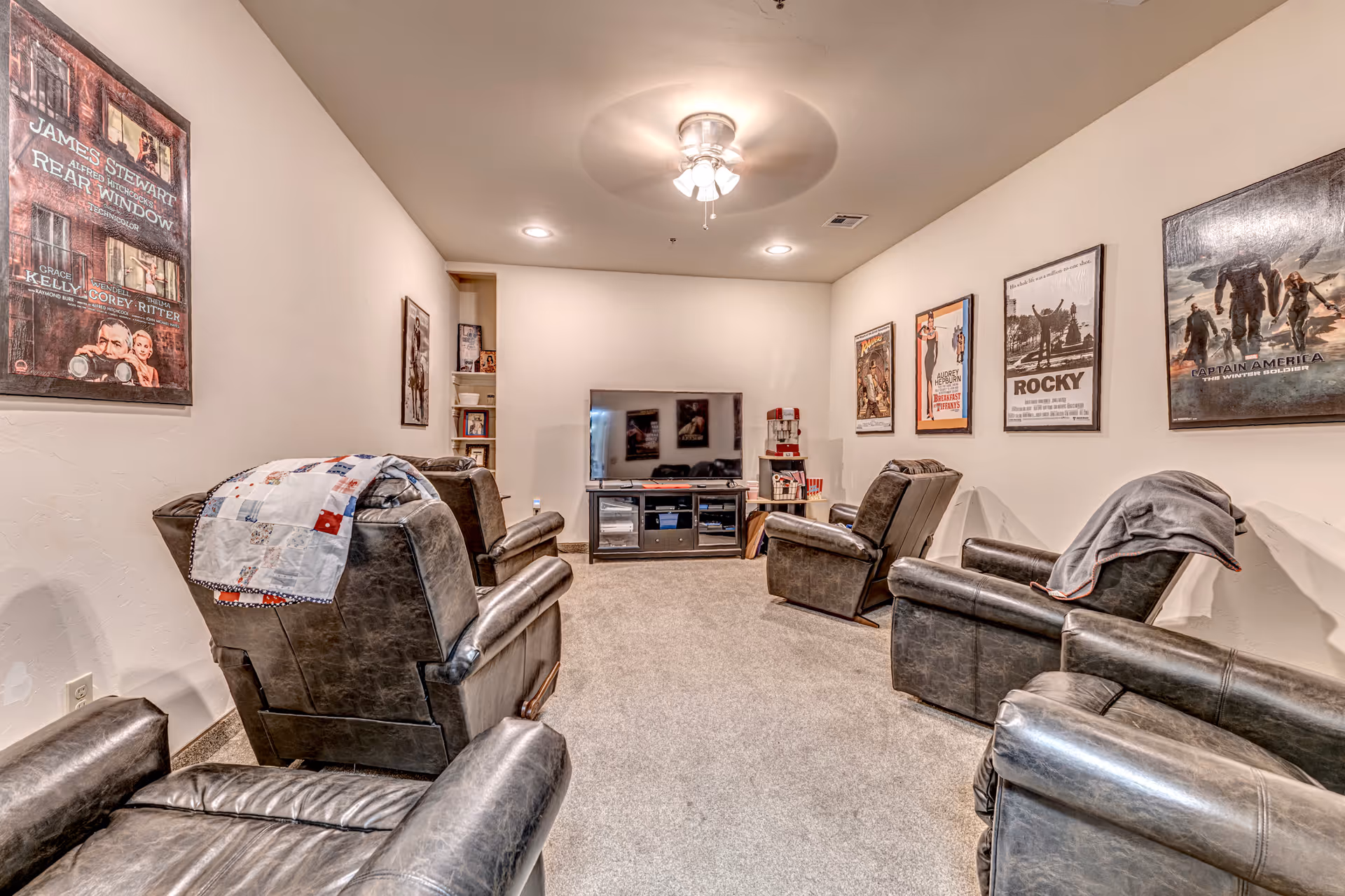 A cozy media room with six leather recliners arranged in two rows facing a flat-screen TV on a stand. The walls are decorated with framed movie posters including Rear Window, Rocky, and Captain America. The room has beige carpet and a ceiling fan with lights.