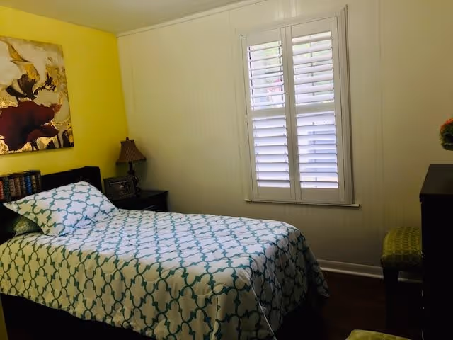 A small bedroom with a single bed covered in a white and teal patterned bedspread. There is a nightstand with a lamp and picture frame next to the bed, a window with white shutters on the wall, and a dresser with a green cushioned chair beside it. The walls are painted yellow and white, and there is a large floral painting hanging above the bed.