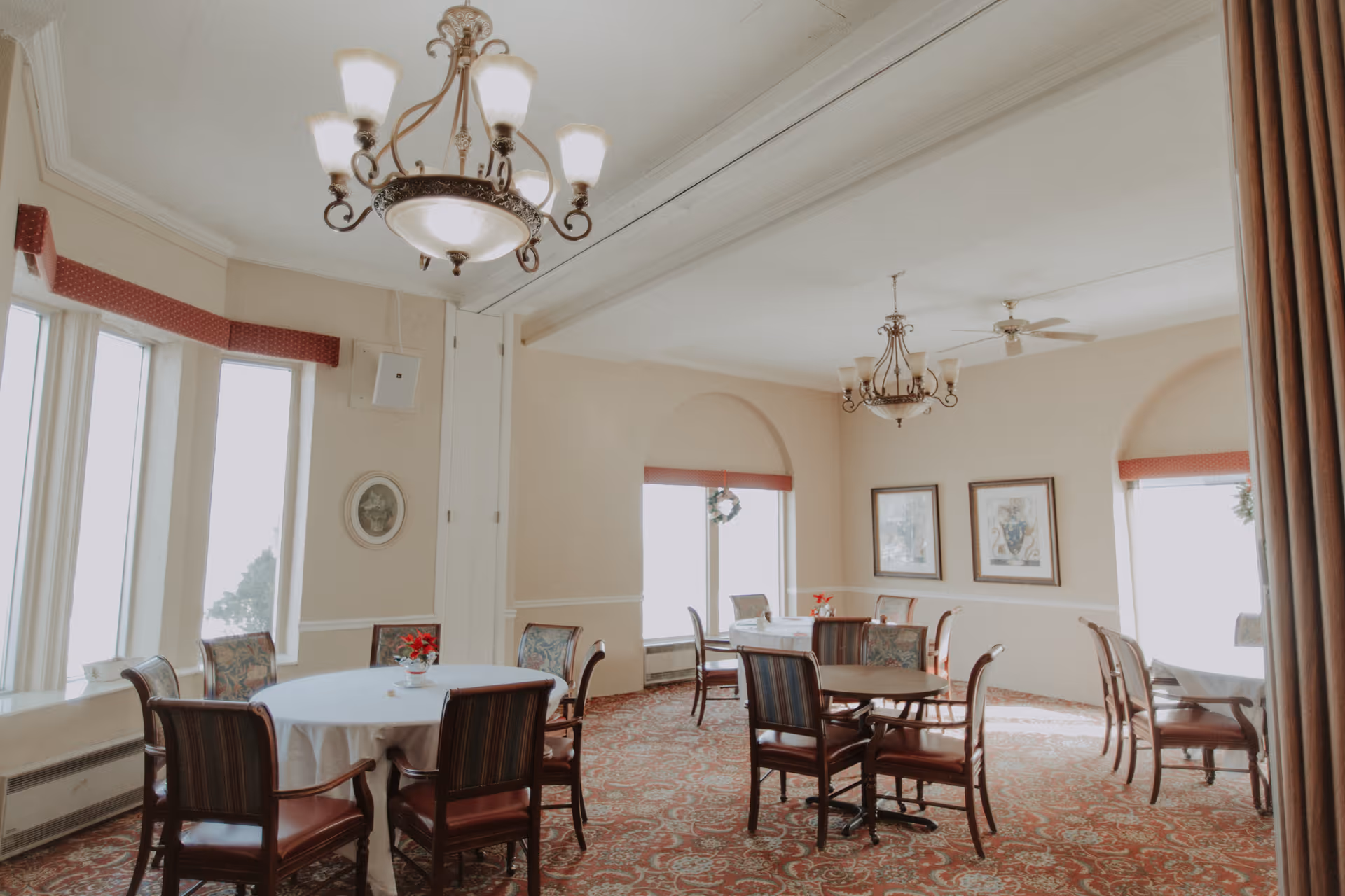A bright dining room with several round tables covered with white tablecloths, surrounded by wooden chairs with patterned upholstery. The room features large windows with red valances, ornate chandeliers, framed artwork on the walls, and a patterned carpet.