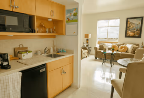 A bright and clean interior space featuring a small kitchen area with wooden cabinets, a microwave, a coffee maker, and a mini refrigerator. Adjacent to the kitchen is a cozy living room with a beige sofa, round glass coffee table, cushioned chairs, a floor lamp, and a large window letting in natural light.