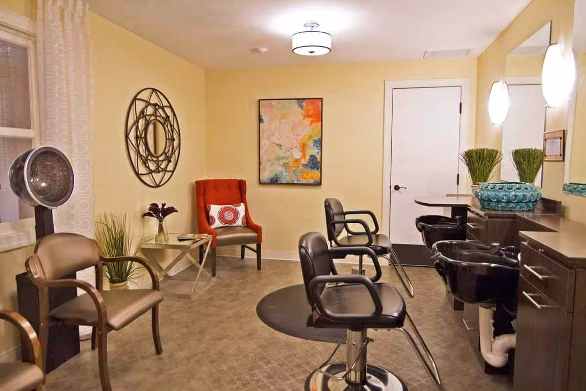 Interior of a salon area in a senior living facility with two black salon chairs in front of wash basins, a red armchair with a decorative pillow, a brown chair, a hair dryer, a small table with plants and flowers, a colorful abstract painting on the wall, and soft yellow walls.