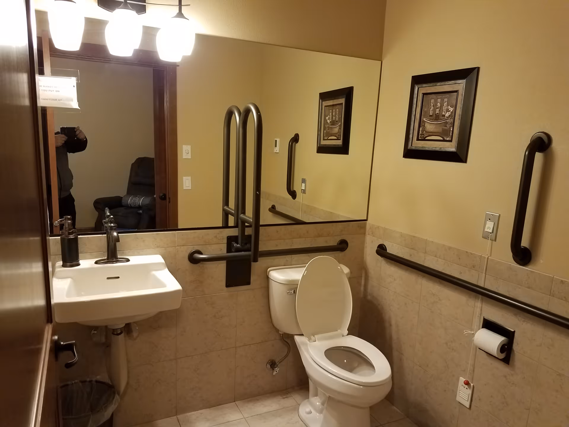 Accessible bathroom with a toilet (seat up), wall-mounted sink, multiple grab bars, a large mirror, and framed artwork.