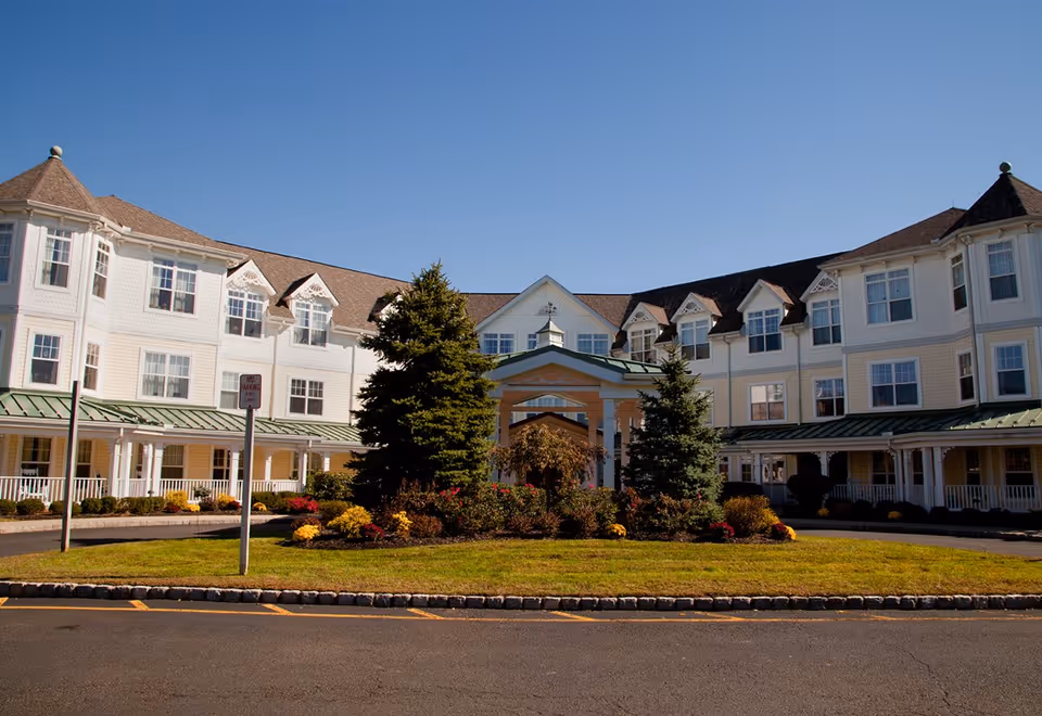 Front view of a large white multi-story senior living building with a covered entrance and landscaped circular driveway under a clear blue sky.