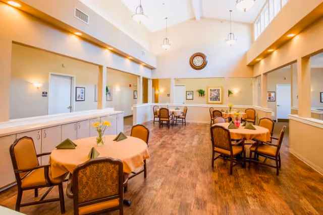 A bright and spacious dining area in a senior living facility with round tables covered in peach-colored tablecloths, each set with folded green napkins and small flower vases. The room features high ceilings with hanging light fixtures, wood flooring, and light-colored walls adorned with framed artwork and a large clock.