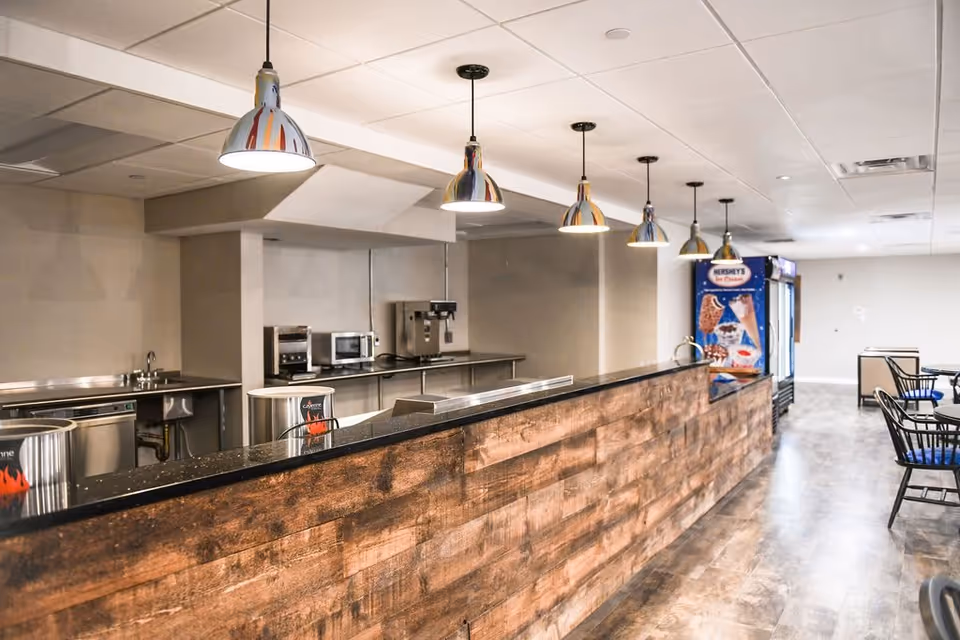Interior view of a modern kitchen and dining area with a long wooden counter, hanging pendant lights, stainless steel appliances including a microwave and coffee machine, and a Hershey's ice cream vending machine in the background. There are tables and chairs visible on the right side.