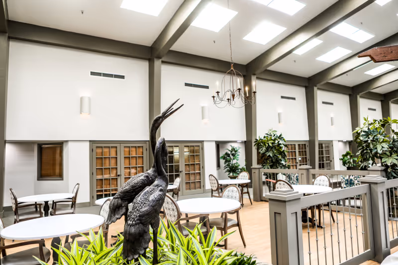 Bright indoor dining atrium with round tables and chairs, skylights, plants, and a decorative heron sculpture.