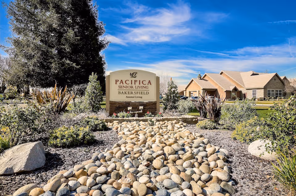 Outdoor landscaped area with a dry riverbed made of smooth stones leading to a sign that reads 'Pacifica Senior Living Bakersfield Assisted Living'. There are bushes, trees, and a building with a tan roof in the background under a blue sky with some clouds.