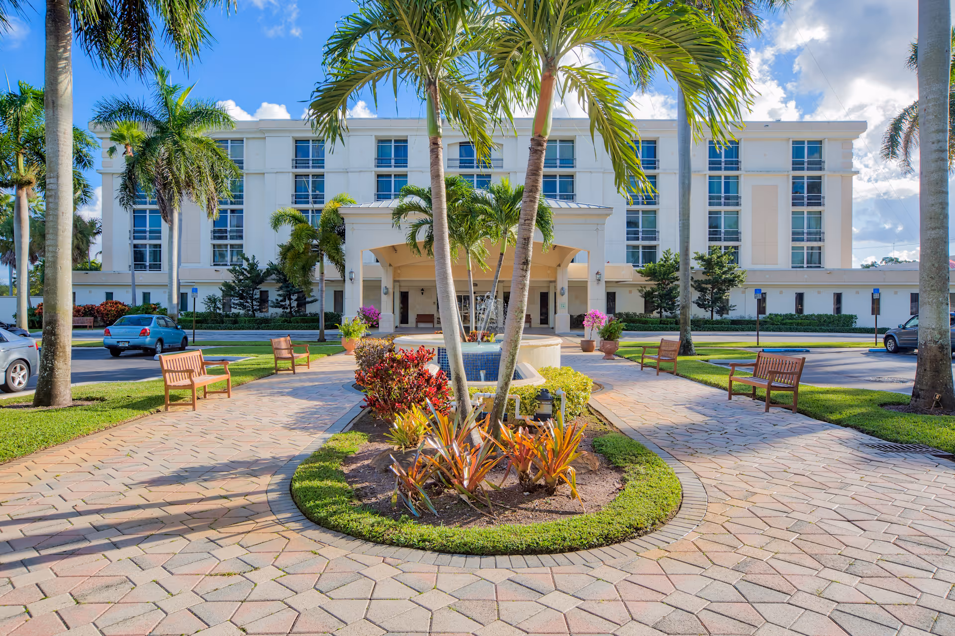 Front exterior view of The Peninsula Assisted Living & Memory Care building with a paved walkway, palm trees, benches, and a central fountain surrounded by landscaped plants under a blue sky with clouds.