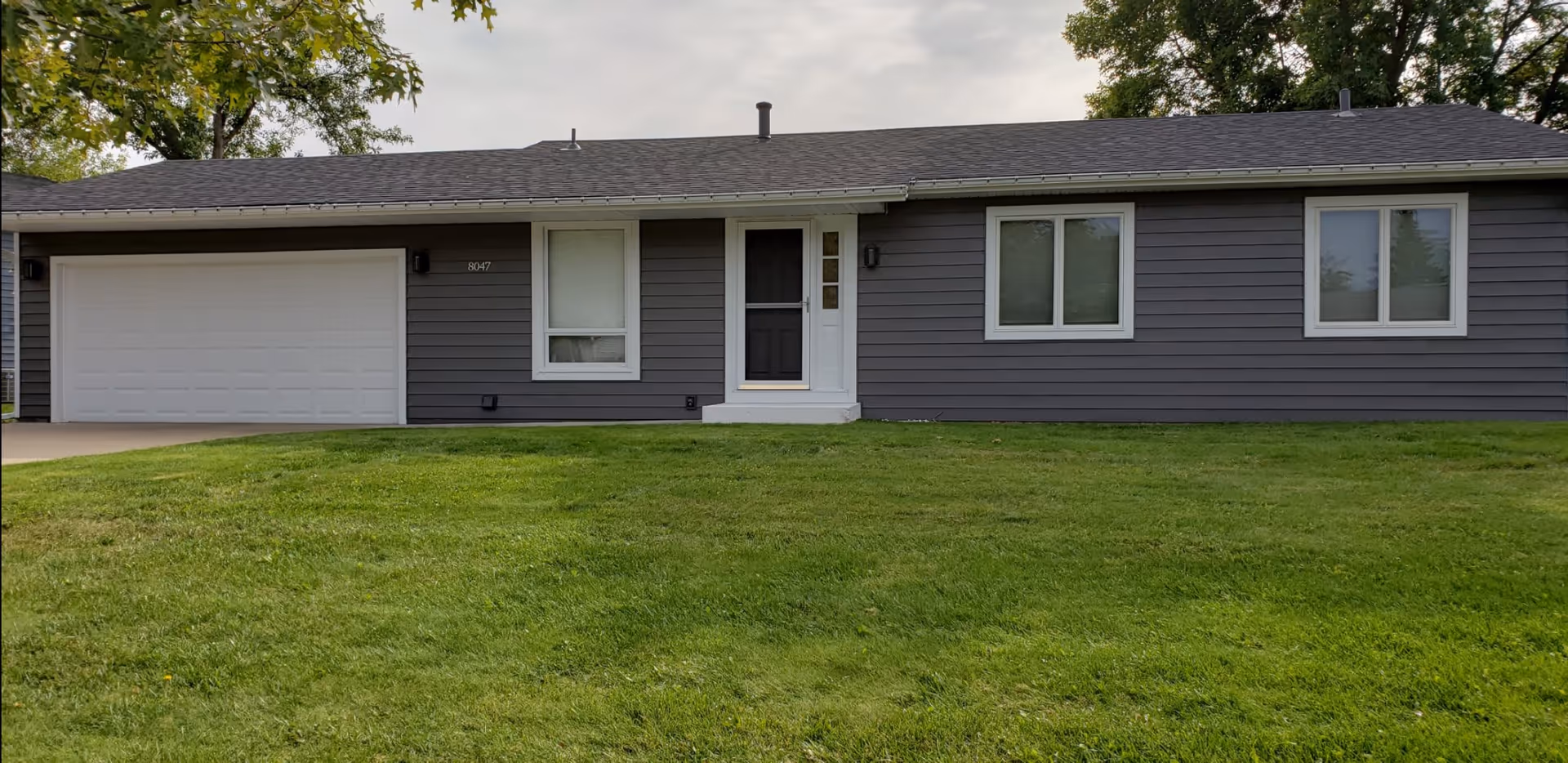 Single-story gray house with a black front door, two white-framed windows, a white garage door, and a well-maintained green lawn in front. Trees are visible in the background.