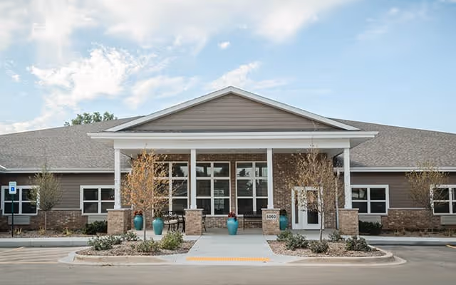 Front exterior view of Kenosha Place Senior Living building with a covered entrance, large windows, and landscaped area with small trees and plants under a partly cloudy sky.
