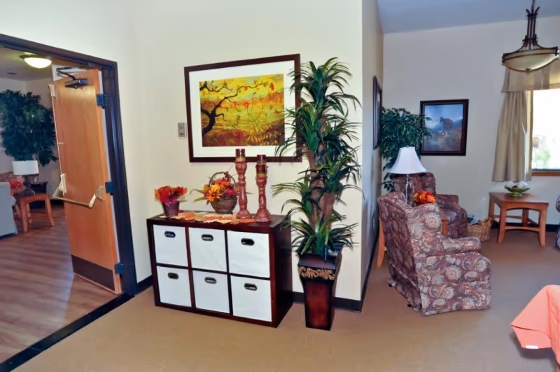Interior view of a senior living facility common area with a storage unit holding decorative items like candles and flowers, a tall potted plant beside it, and a seating area with two patterned armchairs, a side table with a lamp, and framed artwork on the walls.