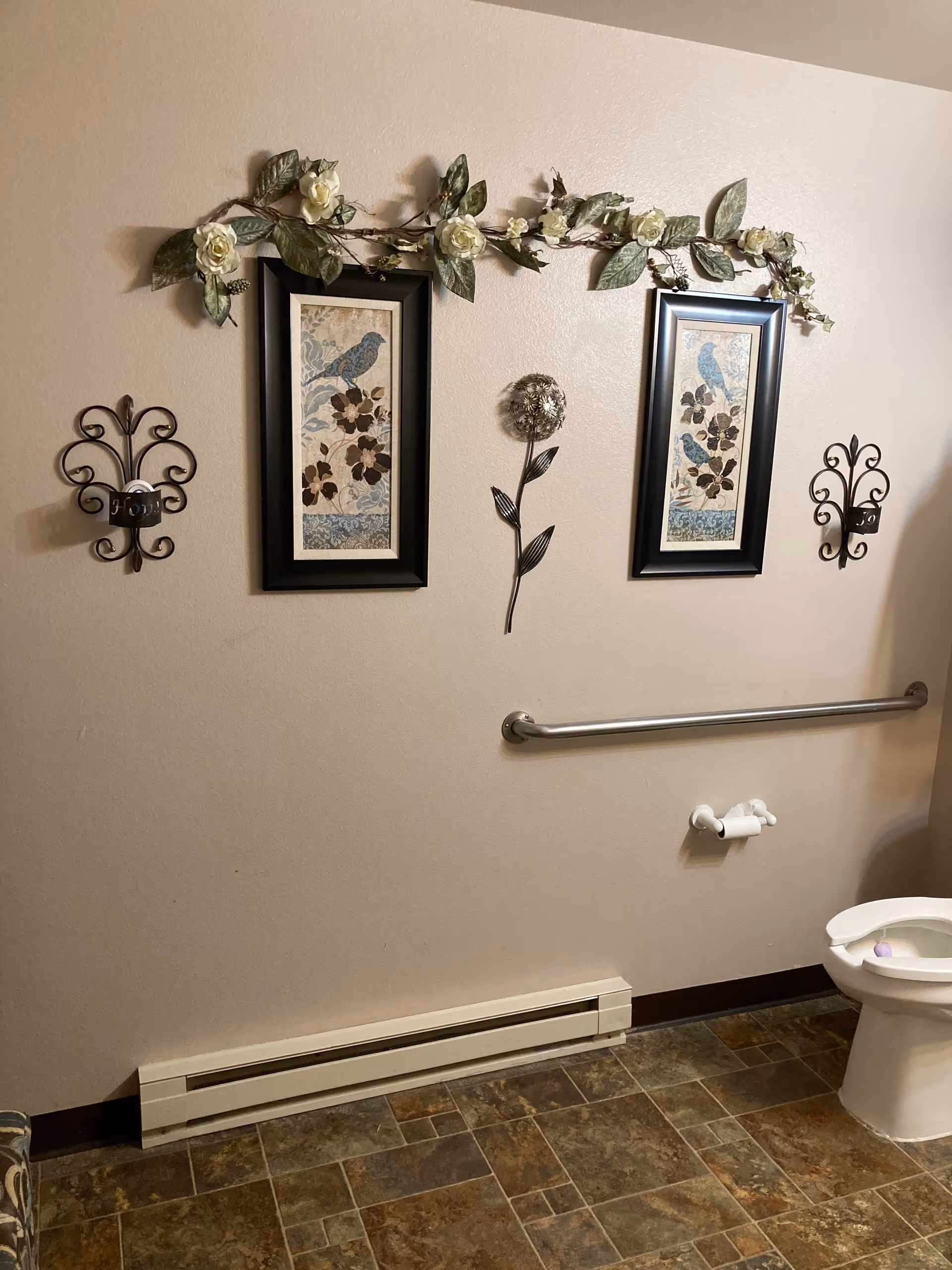 A bathroom wall decorated with two framed pictures of birds and flowers, a metal flower wall art piece in the center, and a garland of white flowers and green leaves draped above. Below the decorations is a metal grab bar, a toilet paper holder with a roll of toilet paper, and part of a white toilet. The floor has brown and tan patterned tiles.