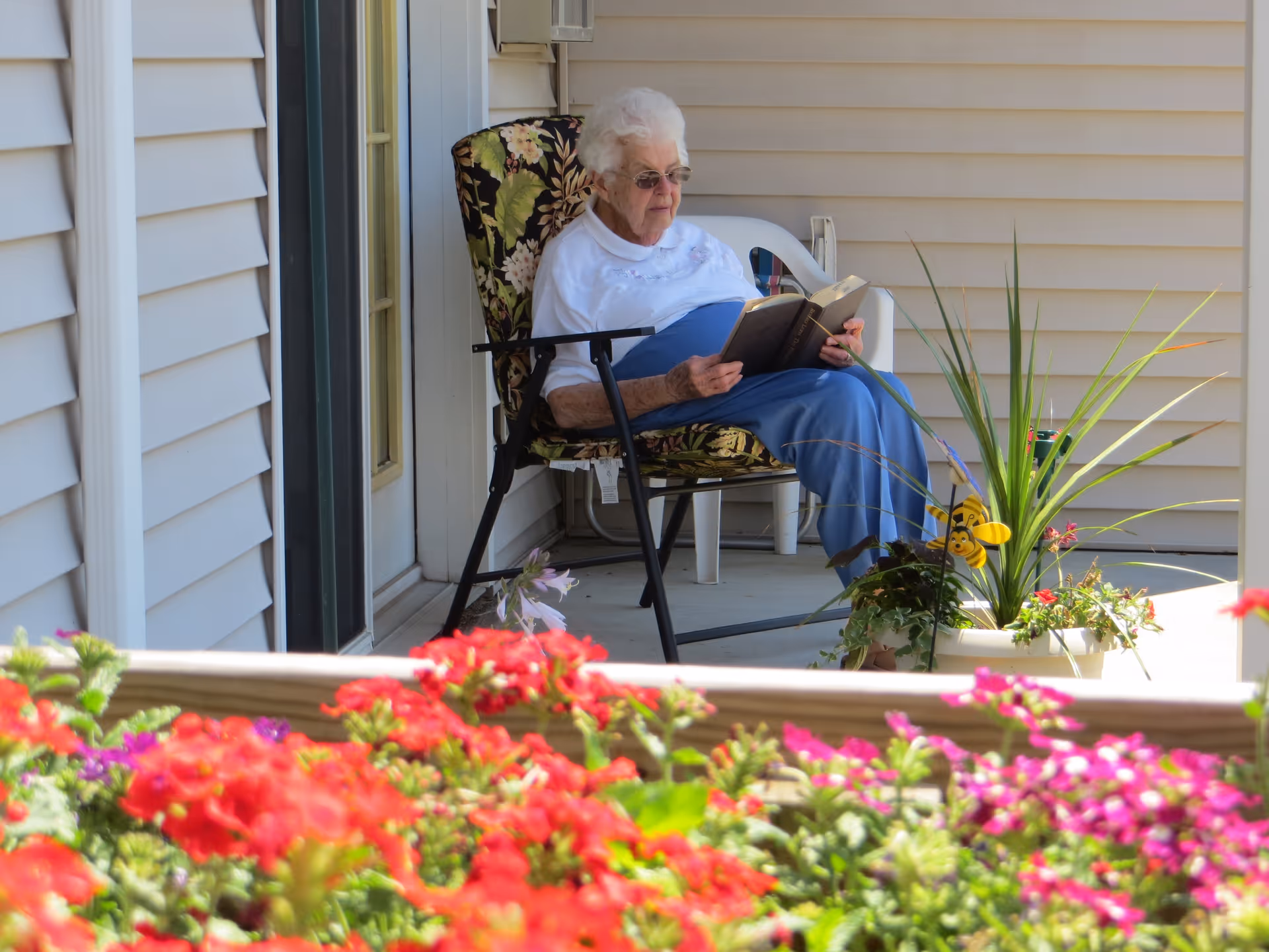An elderly woman with white hair and glasses sitting on a floral cushioned chair on a porch, reading a book. There are colorful flowers in the foreground and a potted plant with a decorative bee on the porch beside her. The porch has light-colored siding and a white plastic chair behind her.