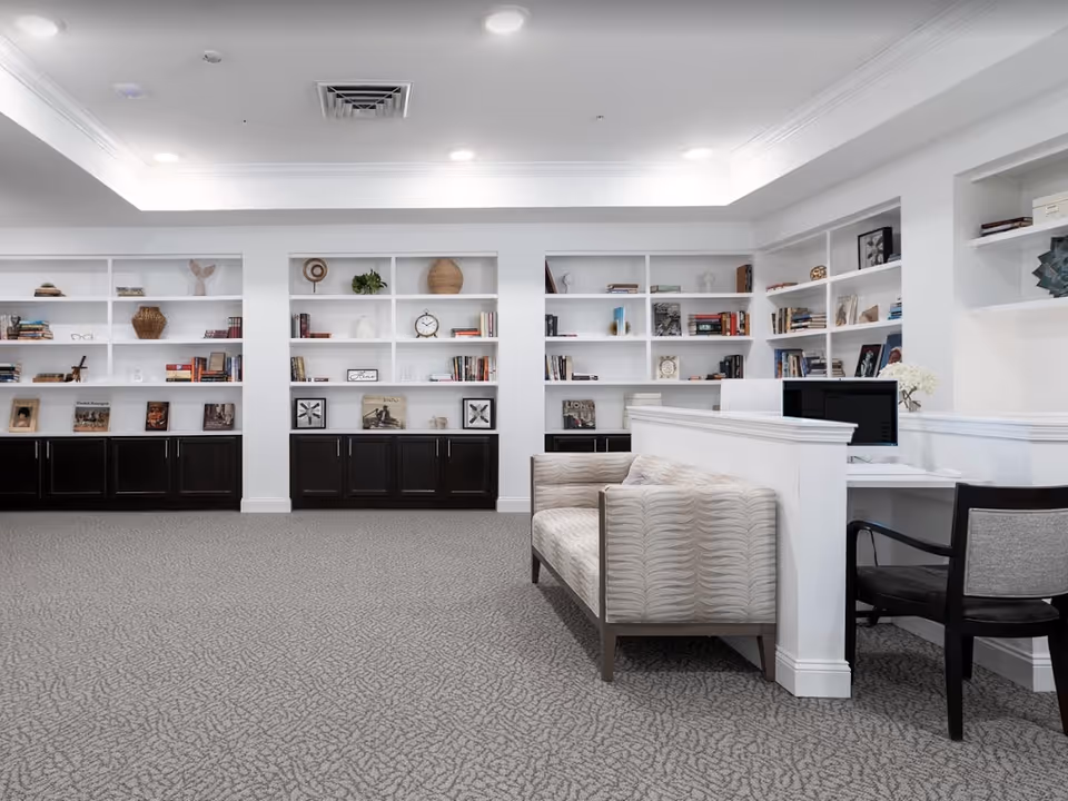 A bright and spacious interior room with white built-in bookshelves filled with books and decorative items. There is a patterned beige sofa and a black chair positioned near a white desk with a computer. The room has a light gray carpet and recessed ceiling lights.