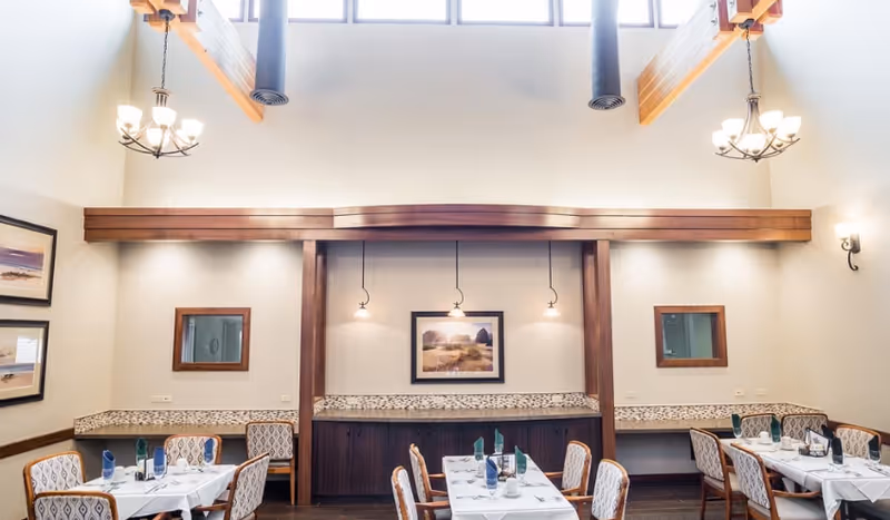 Dining room with set tables and upholstered chairs, chandeliers, high ceilings, and framed artwork on a wood-accented wall.