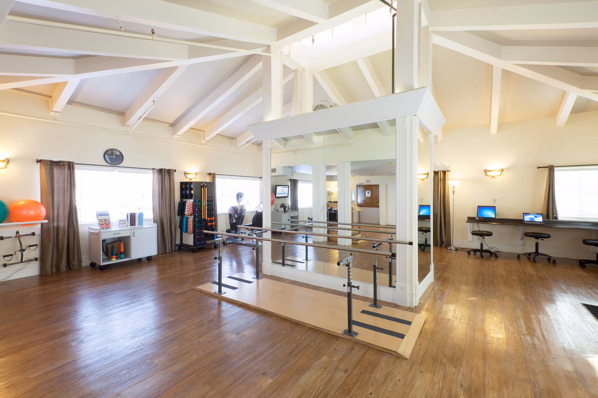 Spacious interior rehabilitation/activity room with parallel walking bars, mirrored columns, exercise equipment, and computer stations.