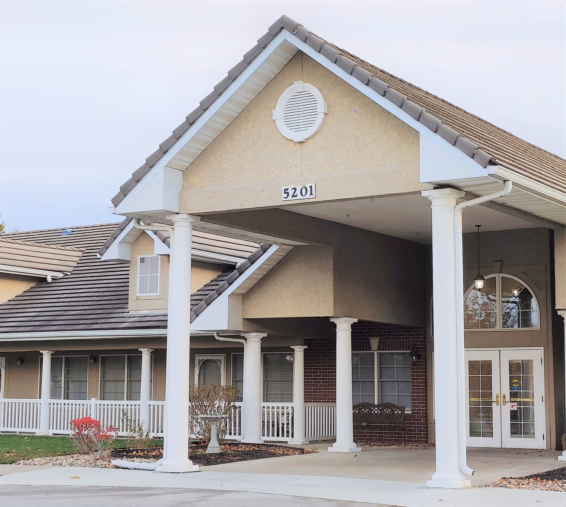 Front exterior view of a senior living facility building with beige walls, white columns, a covered entrance, and the number 5201 displayed above the entrance.