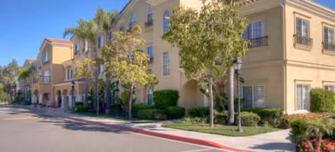 Sunlit beige senior living facility with arches, trees, and a quiet street