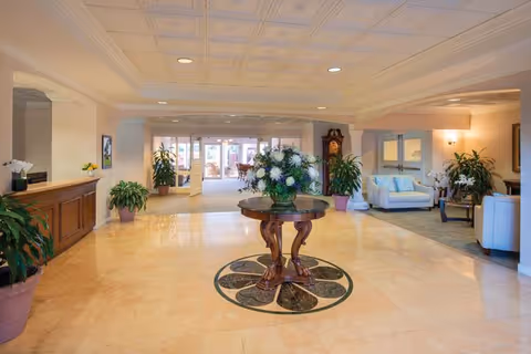Spacious and well-lit lobby area with a round wooden table featuring a large floral arrangement in the center. The floor is tiled with a decorative circular inlay beneath the table. There are several potted plants placed around the room, a wooden reception desk on the left, and comfortable seating areas with sofas and chairs on the right. Large windows and glass doors are visible at the far end, allowing natural light to fill the space.