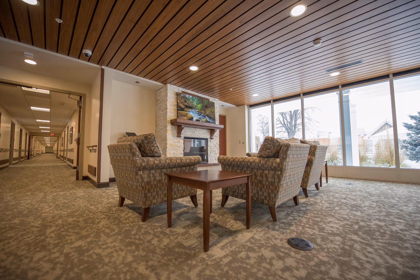 A cozy seating area in a senior living facility with four patterned armchairs arranged around a wooden coffee table. Behind the seating area is a stone fireplace with a nature-themed painting above it. Large windows on the right side let in natural light and show an outdoor view with trees and snow. A long hallway extends to the left with handrails along the walls and ceiling lights.
