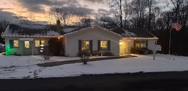 A single-story building covered with snow at dusk, decorated with holiday lights along the roofline and around the entrance. There is a small pathway leading to the front door, a bench under the windows, and an American flag on a pole to the right. Trees without leaves are visible in the background under a cloudy sky.