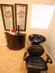 A small corner of a room featuring a black salon chair with a black wash basin behind it. Next to the chair is a dark wooden cabinet with two framed floral artworks hanging above it. A window with white blinds lets in natural light, and the floor is tiled in a light beige color.