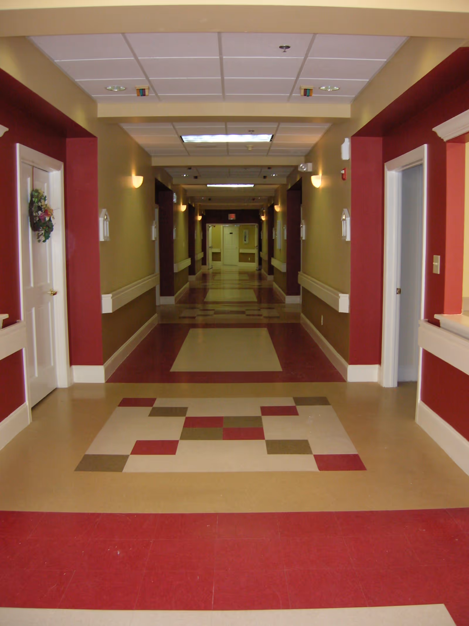 A long, well-lit hallway in a senior living facility with beige and burgundy walls and patterned floor tiles. Doors line both sides of the hallway, and there are wall-mounted lights and handrails along the walls.
