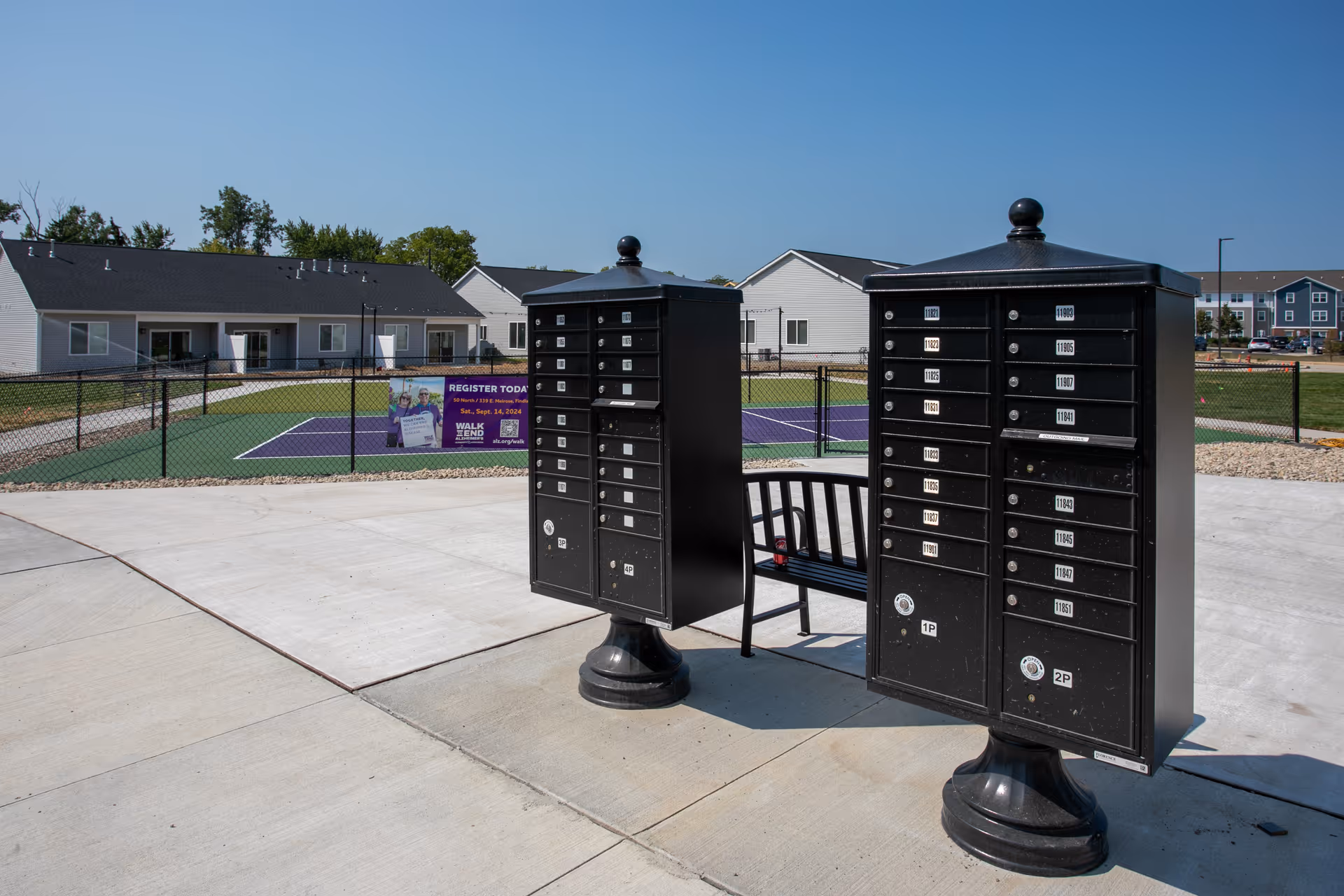 Two black community mailbox units on a paved area with a fenced tennis court and apartment buildings in the background.