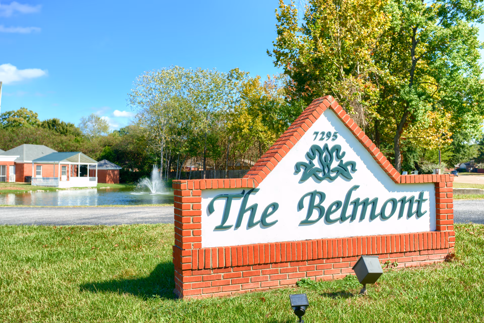 Outdoor view of a brick and white sign with the text 'The Belmont' and the number 7295, set on a grassy area with trees, a pond with a fountain, and buildings in the background under a clear blue sky.