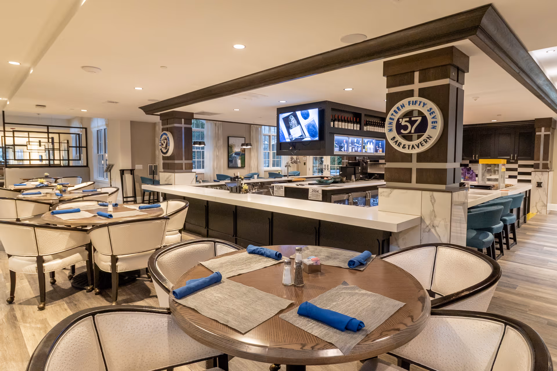 Modern dining room with round tables set with blue napkins and a central bar area featuring a '57' sign.