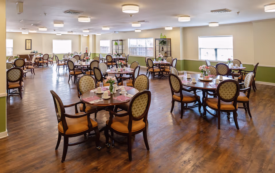 A spacious dining room with multiple round wooden tables, each surrounded by cushioned chairs with patterned backs. The tables are set with placemats, cups, glasses, and small flower arrangements. The room has wooden flooring, light-colored walls with a green lower half, and several windows letting in natural light. Ceiling lights provide additional illumination.