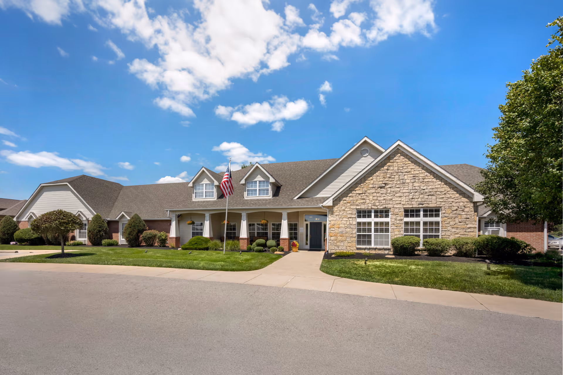 Exterior view of a single-story senior living facility building with a combination of stone and siding facade, a front porch with columns, an American flag on a flagpole, well-maintained green lawn, shrubs, and a clear blue sky with some clouds.