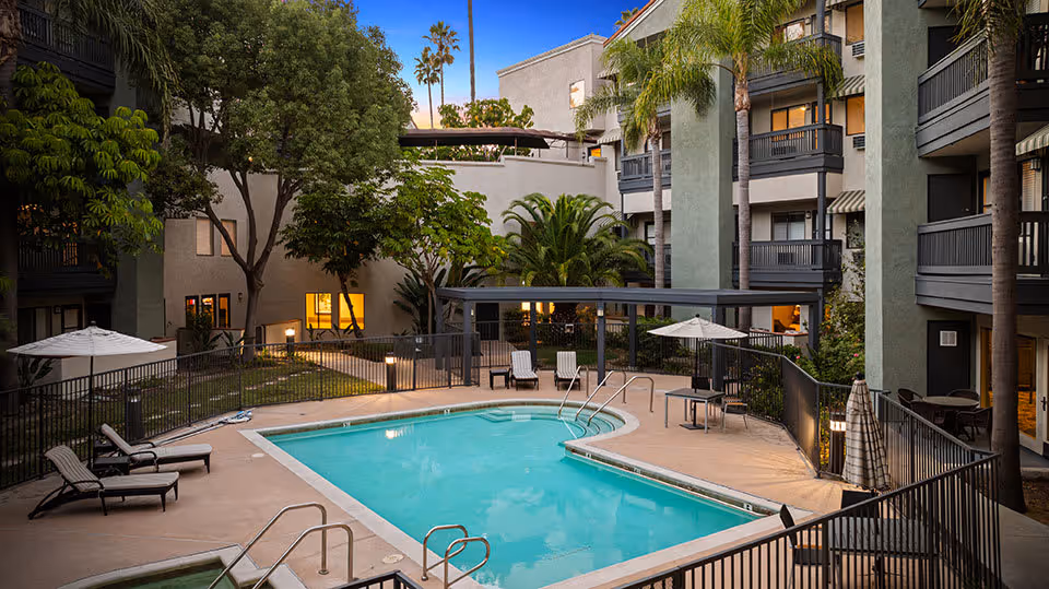 Outdoor swimming pool area at Ivy Park of Wellington, surrounded by a black metal fence with lounge chairs and umbrellas. The pool is situated in the courtyard of a multi-story residential building with balconies and palm trees, under a clear evening sky.