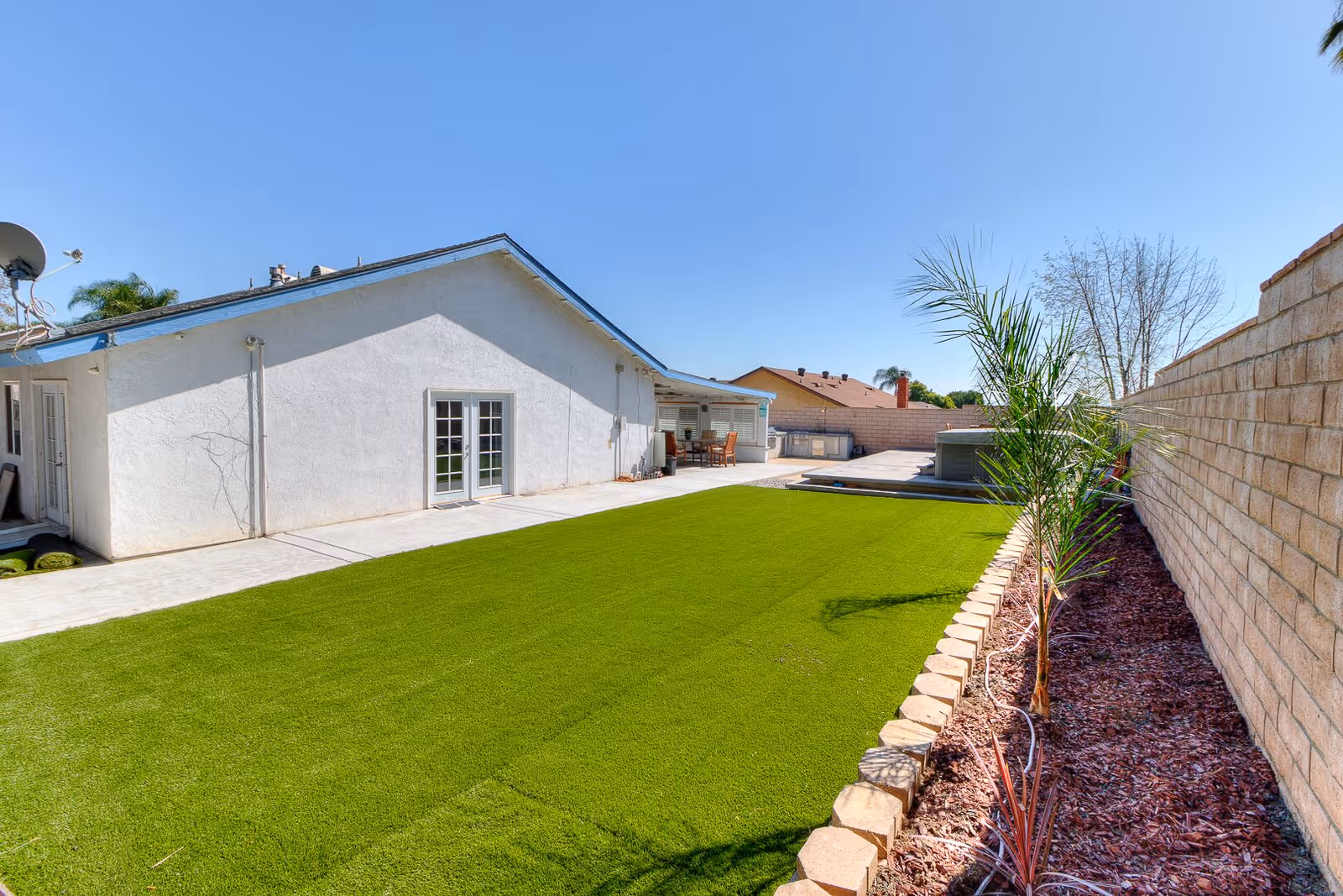 Outdoor view of a backyard area at Bella Maison Assisted Living featuring a well-maintained green lawn, a white building with French doors, a patio area with outdoor furniture, a hot tub, and a brick wall with small plants along the edge under a clear blue sky.