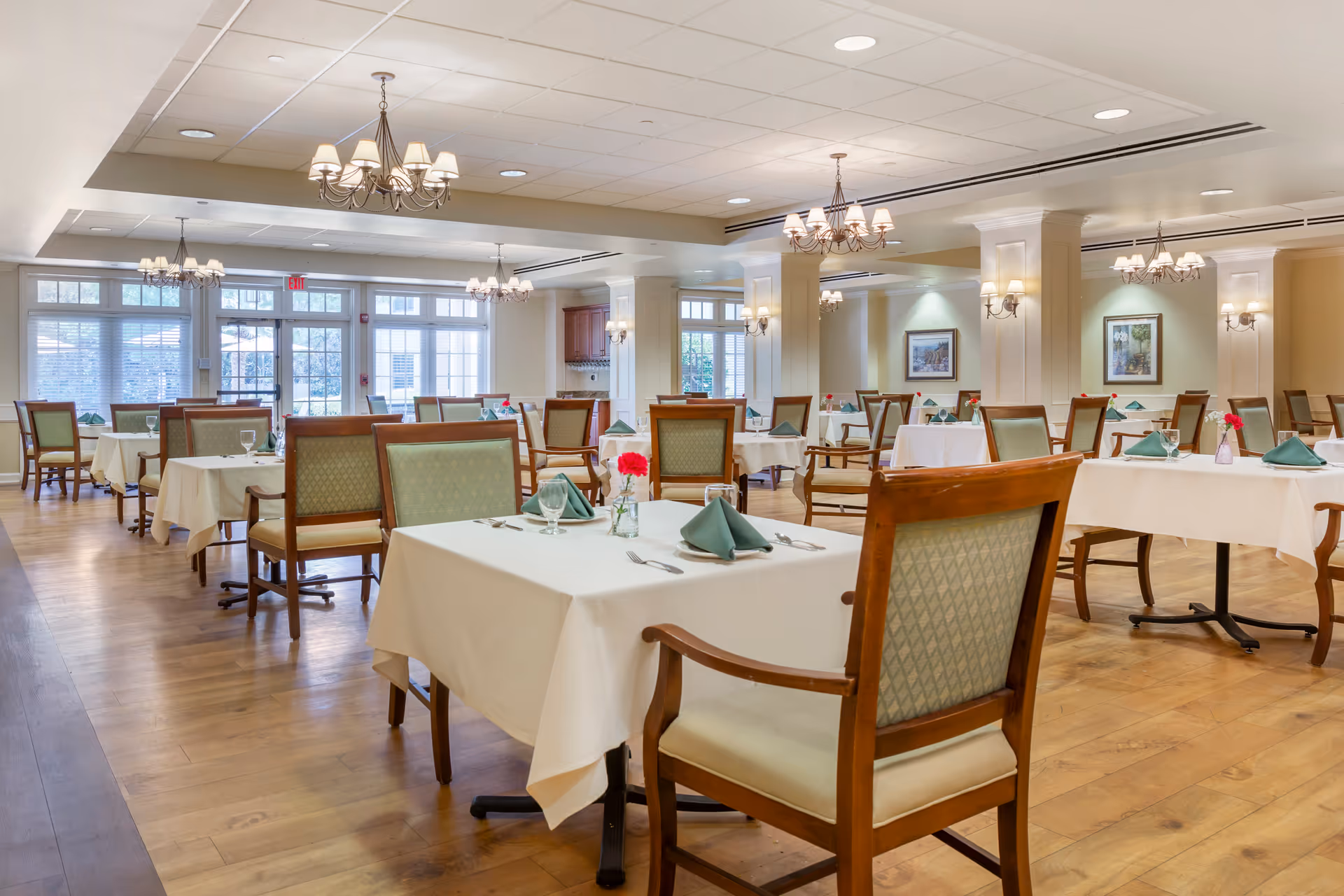 Bright, spacious dining room with neatly set tables, wooden chairs, chandeliers, and hardwood floors.